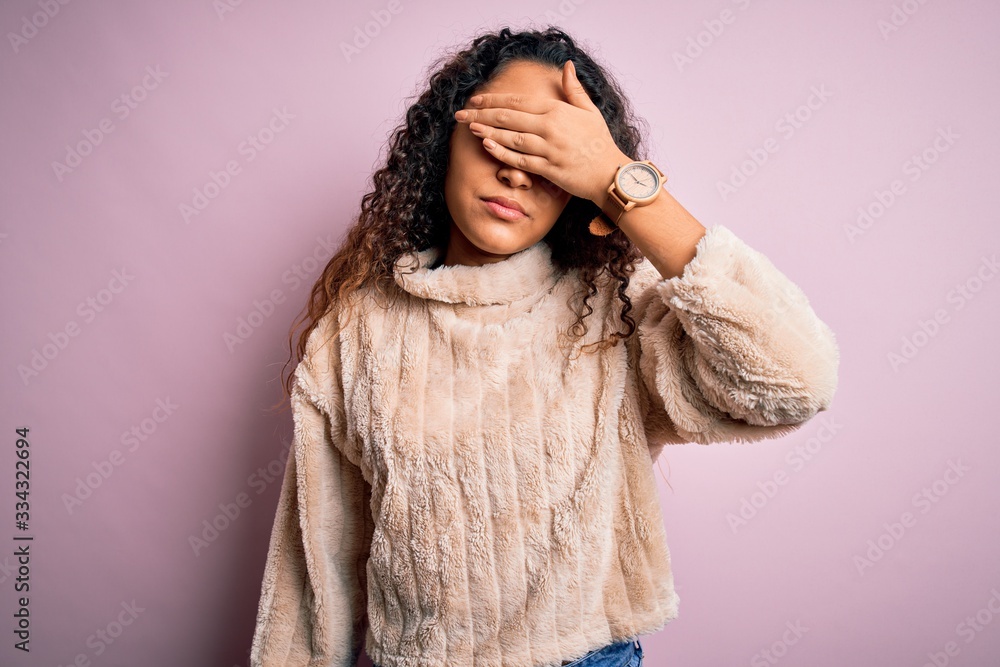 Young beautiful woman with curly hair wearing casual sweater standing over pink background covering eyes with hand, looking serious and sad. Sightless, hiding and rejection concept Young beautiful woman with curly hair wearing casual sweater standing over pink background covering eyes with hand, looking serious and sad. Sightless, hiding and rejection concept
