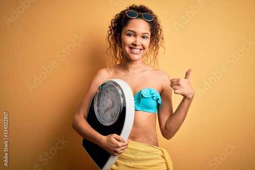 Young beautiful american slim woman on vacation wearing bikini holding weight machine happy with big smile doing ok sign  thumb up with fingers  excellent sign