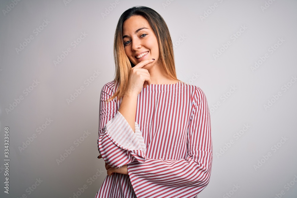 Young beautiful blonde woman with blue eyes wearing stiped t-shirt over white background looking confident at the camera with smile with crossed arms and hand raised on chin. Thinking positive. Young beautiful blonde woman with blue eyes wearing stiped t-shirt over white background looking confident at the camera with smile with crossed arms and hand raised on chin. Thinking positive.