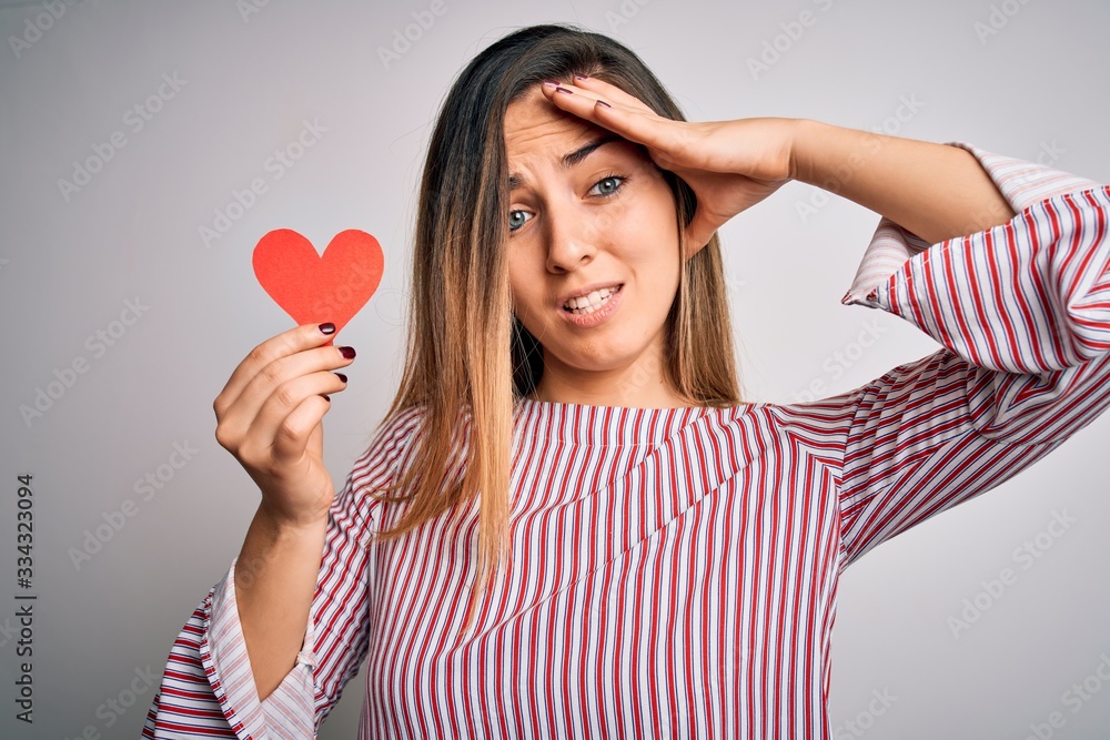 Young beautiful blonde romantic woman holding red paper heart over white background stressed with hand on head, shocked with shame and surprise face, angry and frustrated. Fear and upset for mistake. Young beautiful blonde romantic woman holding red paper heart over white background stressed with hand on head, shocked with shame and surprise face, angry and frustrated. Fear and upset for mistake.