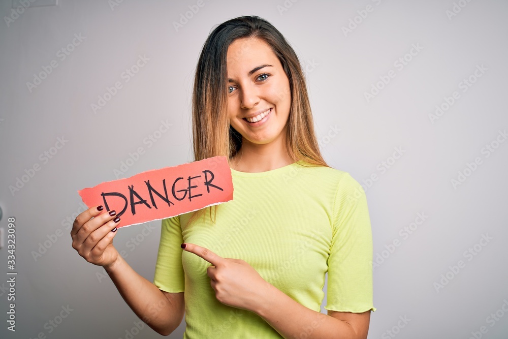 Young beautiful blonde woman with blue eyes holding paper with danger message very happy pointing with hand and finger Young beautiful blonde woman with blue eyes holding paper with danger message very happy pointing with hand and finger