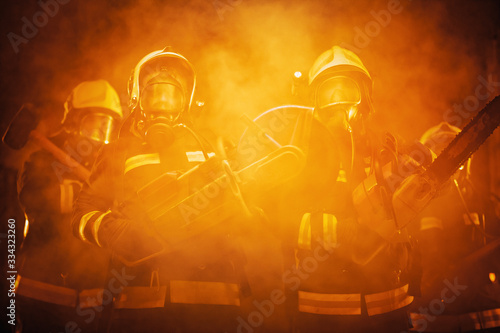 Group of professional firefighters wearing full equipment  oxygen masks  and emergency rescue tools  circular hydraulic and gas saw  axe  and sledge hammer. smoke and fire trucks in the background.