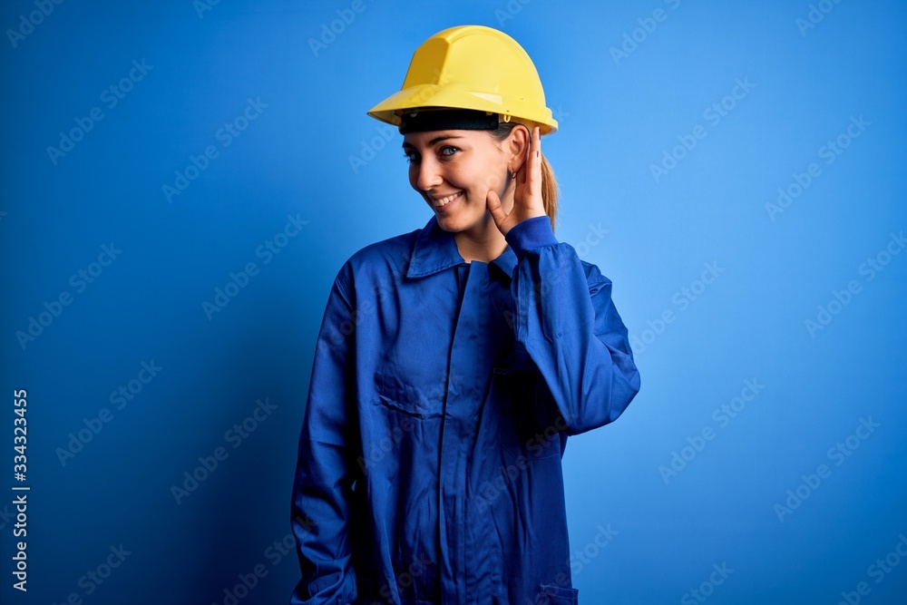 Young beautiful worker woman with blue eyes wearing security helmet and uniform smiling with hand over ear listening an hearing to rumor or gossip. Deafness concept.