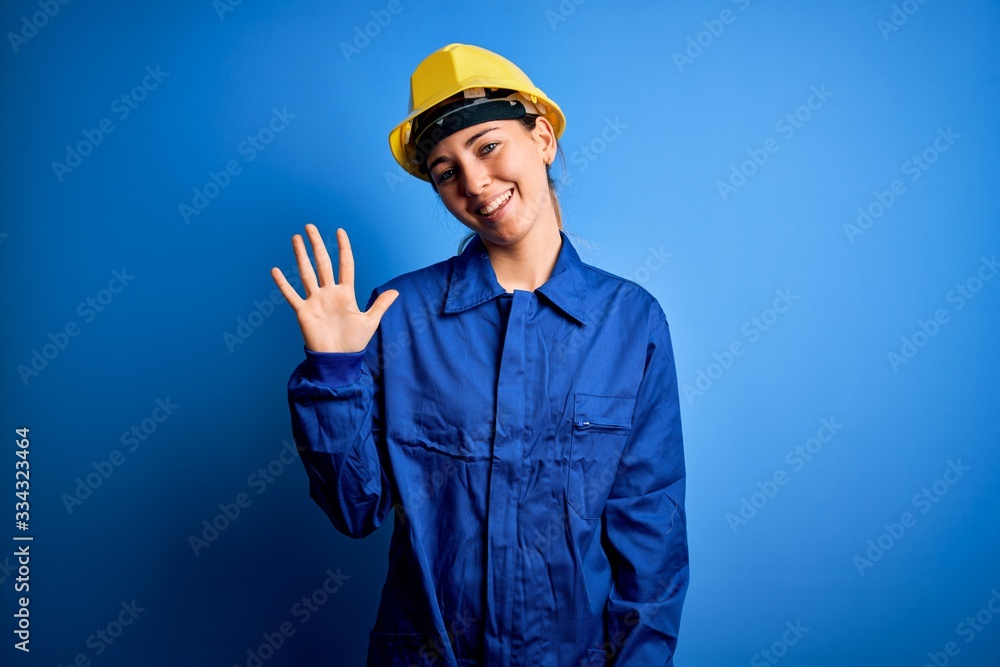 Young beautiful worker woman with blue eyes wearing security helmet and uniform showing and pointing up with fingers number five while smiling confident and happy. Young beautiful worker woman with blue eyes wearing security helmet and uniform showing and pointing up with fingers number five while smiling confident and happy.