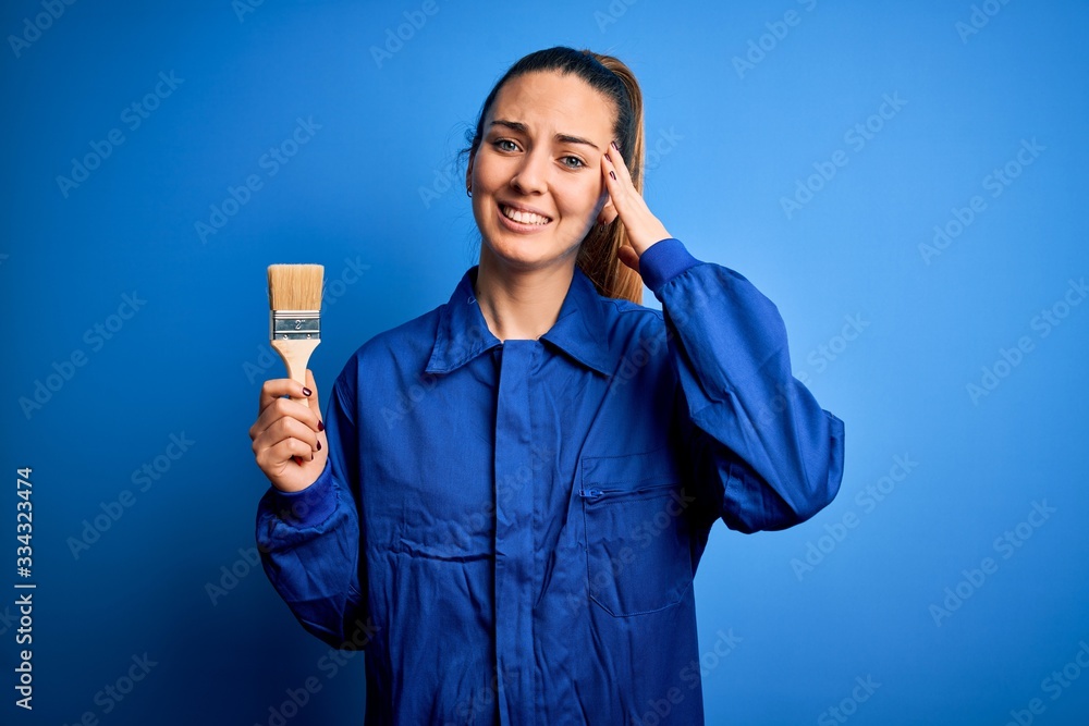 Young beautiful painter woman with blue eyes painting wearing uniform using paint brush stressed with hand on head, shocked with shame and surprise face, angry and frustrated. Fear and upset Young beautiful painter woman with blue eyes painting wearing uniform using paint brush stressed with hand on head, shocked with shame and surprise face, angry and frustrated. Fear and upset