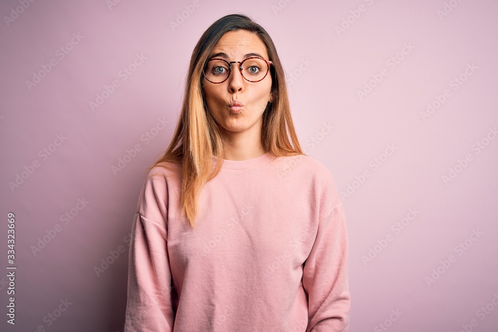Beautiful blonde woman with blue eyes wearing sweater and glasses over pink background making fish face with lips, crazy and comical gesture. Funny expression. Beautiful blonde woman with blue eyes wearing sweater and glasses over pink background making fish face with lips, crazy and comical gesture. Funny expression.