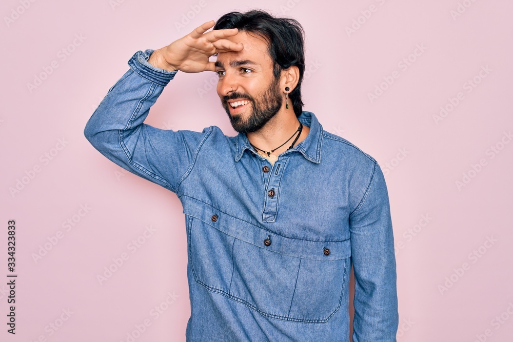 Young handsome hispanic bohemian man wearing hippie style over pink background very happy and smiling looking far away with hand over head. Searching concept. Young handsome hispanic bohemian man wearing hippie style over pink background very happy and smiling looking far away with hand over head. Searching concept.