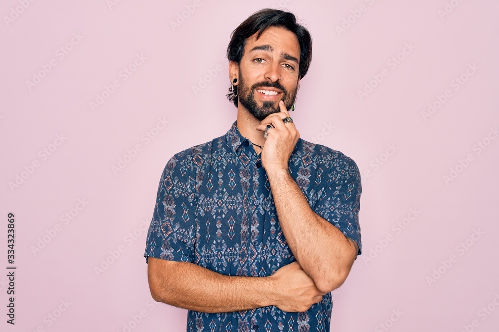 Young handsome hispanic bohemian man wearing hippie style over pink background looking confident at the camera smiling with crossed arms and hand raised on chin. Thinking positive.