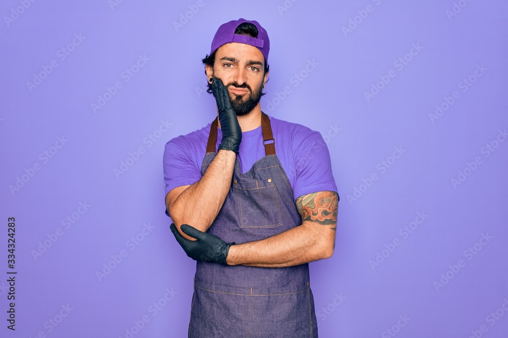 Young handsome hispanic man with bear wearing professional apron working as tattoo artist thinking looking tired and bored with depression problems with crossed arms. Young handsome hispanic man with bear wearing professional apron working as tattoo artist thinking looking tired and bored with depression problems with crossed arms.