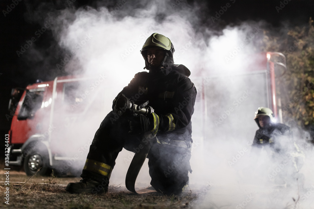 Team of professional firefighters holding water hose in front of a firetruck with smoke in the air. Half silhouette. Team of professional firefighters holding water hose in front of a firetruck with smoke in the air. Half silhouette.