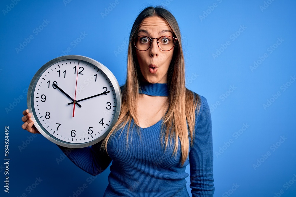 Young blonde woman with blue eyes holding big minute clock over isolated background scared in shock with a surprise face, afraid and excited with fear expression Young blonde woman with blue eyes holding big minute clock over isolated background scared in shock with a surprise face, afraid and excited with fear expression