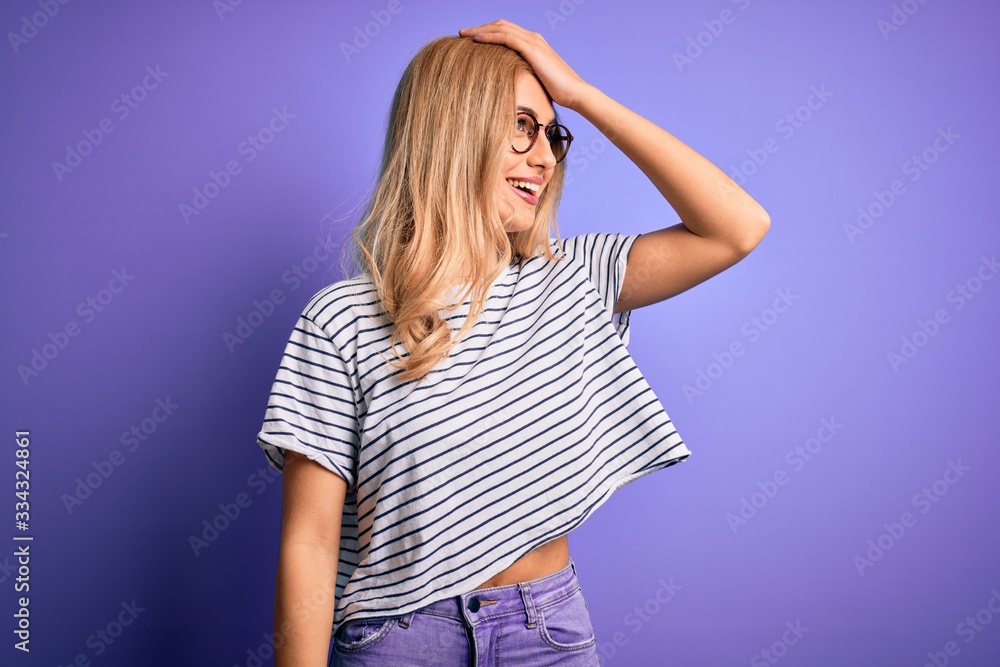 Young beautiful blonde woman wearing striped t-shirt and glasses over purple background smiling confident touching hair with hand up gesture, posing attractive and fashionable Young beautiful blonde woman wearing striped t-shirt and glasses over purple background smiling confident touching hair with hand up gesture, posing attractive and fashionable