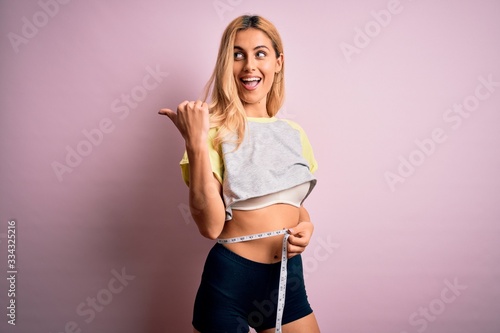 Young beautiful blonde slim woman doing sport using tape measure controlling weight pointing and showing with thumb up to the side with happy face smiling © Krakenimages.com