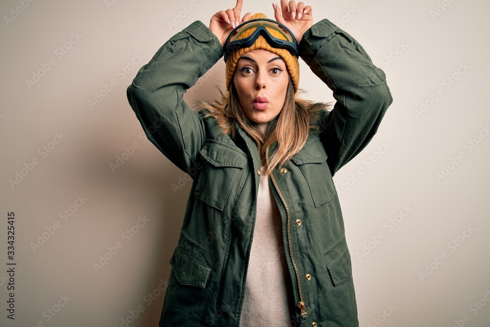 Young brunette skier woman wearing snow clothes and ski goggles over white background doing funny gesture with finger over head as bull horns Young brunette skier woman wearing snow clothes and ski goggles over white background doing funny gesture with finger over head as bull horns
