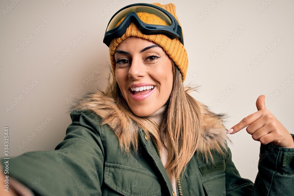 Young brunette skier woman wearing snow clothes and ski goggles make selfie by camera with surprise face pointing finger to himself Young brunette skier woman wearing snow clothes and ski goggles make selfie by camera with surprise face pointing finger to himself