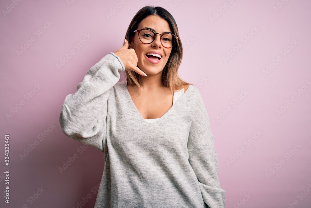Young beautiful brunette woman wearing casual sweater and glasses over pink background smiling doing phone gesture with hand and fingers like talking on the telephone. Communicating concepts. Young beautiful brunette woman wearing casual sweater and glasses over pink background smiling doing phone gesture with hand and fingers like talking on the telephone. Communicating concepts.