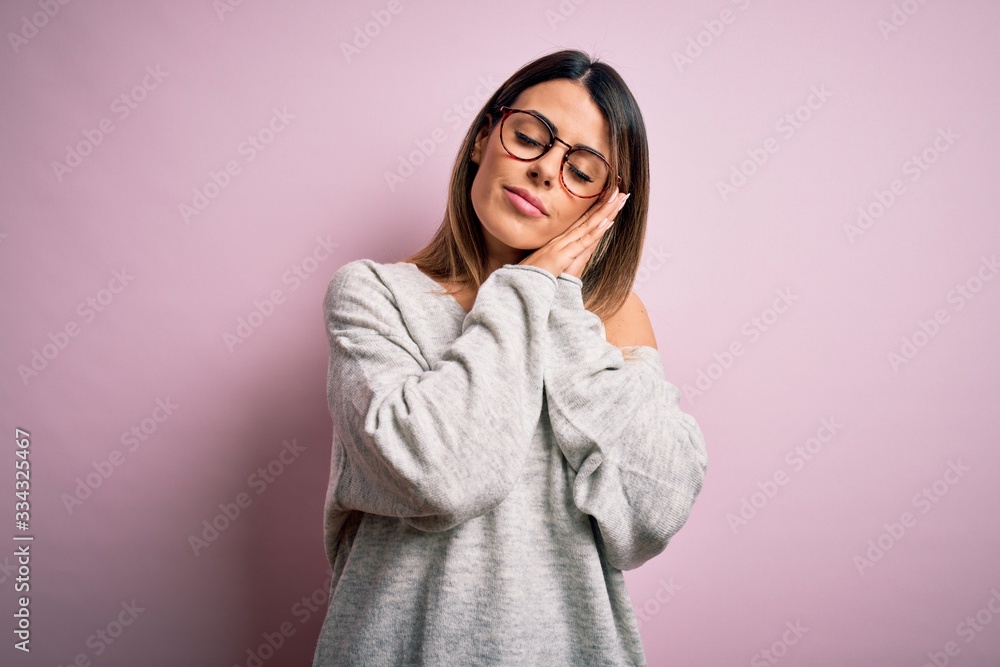 Young beautiful brunette woman wearing casual sweater and glasses over pink background sleeping tired dreaming and posing with hands together while smiling with closed eyes.