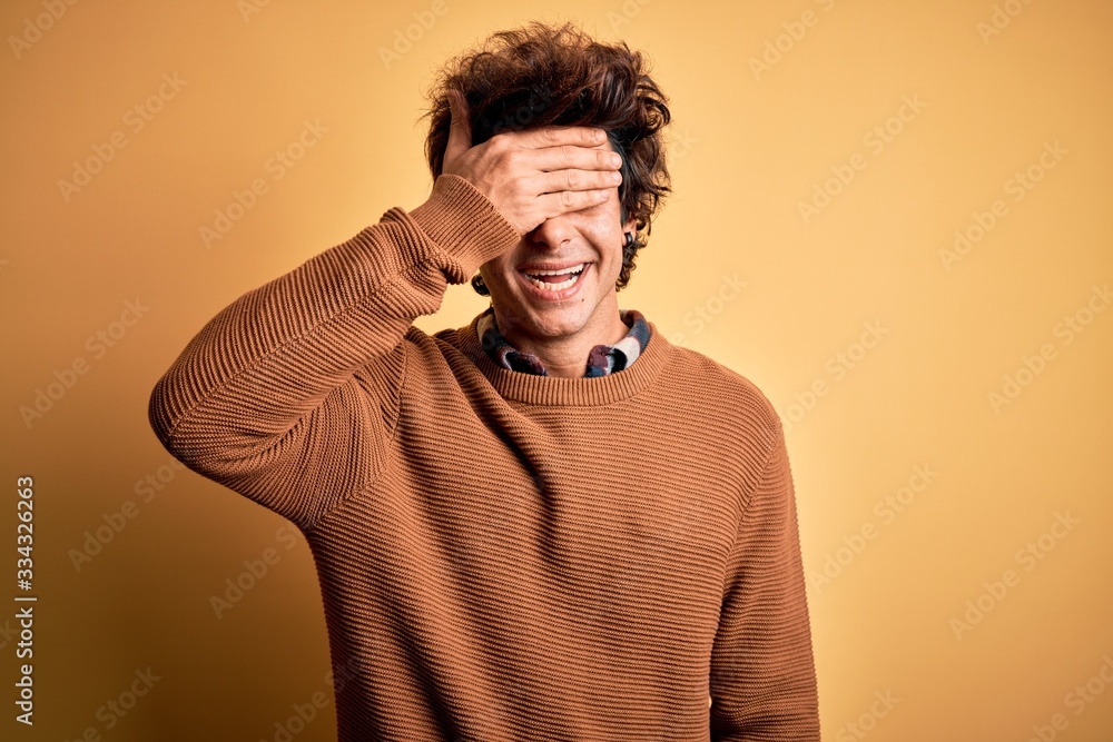 Young handsome man wearing casual shirt and sweater over isolated yellow background smiling and laughing with hand on face covering eyes for surprise. Blind concept. Young handsome man wearing casual shirt and sweater over isolated yellow background smiling and laughing with hand on face covering eyes for surprise. Blind concept.