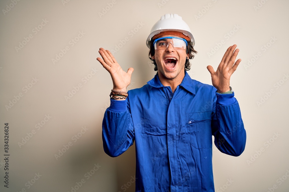 Young constructor man wearing uniform and security helmet over isolated white background celebrating crazy and amazed for success with arms raised and open eyes screaming excited. Winner concept
