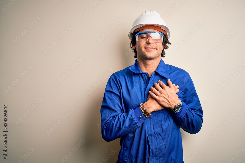 Young constructor man wearing uniform and security helmet over isolated white background smiling with hands on chest with closed eyes and grateful gesture on face. Health concept.