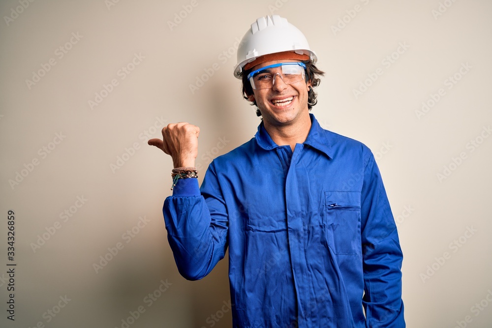 Young constructor man wearing uniform and security helmet over isolated white background smiling with happy face looking and pointing to the side with thumb up. Young constructor man wearing uniform and security helmet over isolated white background smiling with happy face looking and pointing to the side with thumb up.