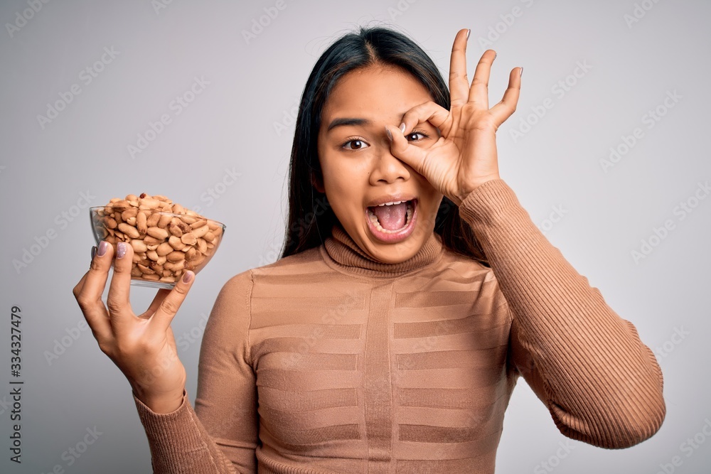 Young asian girl holding bowl of healthy peanuts eating as snack for diet over white background with happy face smiling doing ok sign with hand on eye looking through fingers Young asian girl holding bowl of healthy peanuts eating as snack for diet over white background with happy face smiling doing ok sign with hand on eye looking through fingers