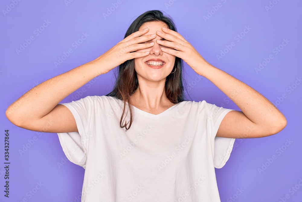 Young beautiful brunette woman wearing casual white t-shirt over purple background covering eyes with hands smiling cheerful and funny. Blind concept. Young beautiful brunette woman wearing casual white t-shirt over purple background covering eyes with hands smiling cheerful and funny. Blind concept.