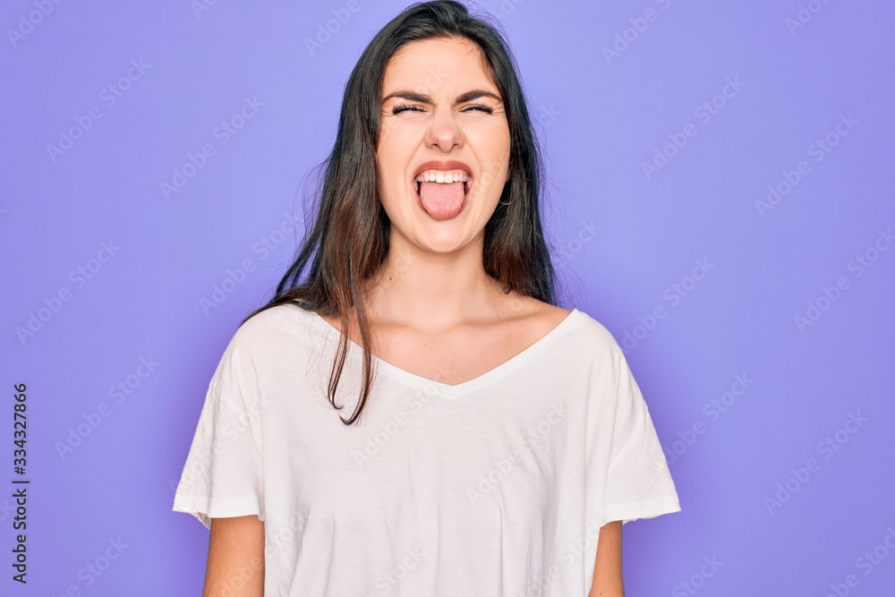 Young beautiful brunette woman wearing casual white t-shirt over purple background sticking tongue out happy with funny expression. Emotion concept. Young beautiful brunette woman wearing casual white t-shirt over purple background sticking tongue out happy with funny expression. Emotion concept.