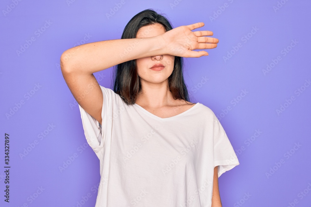 Young beautiful brunette woman wearing casual white t-shirt over purple background covering eyes with arm, looking serious and sad. Sightless, hiding and rejection concept Young beautiful brunette woman wearing casual white t-shirt over purple background covering eyes with arm, looking serious and sad. Sightless, hiding and rejection concept