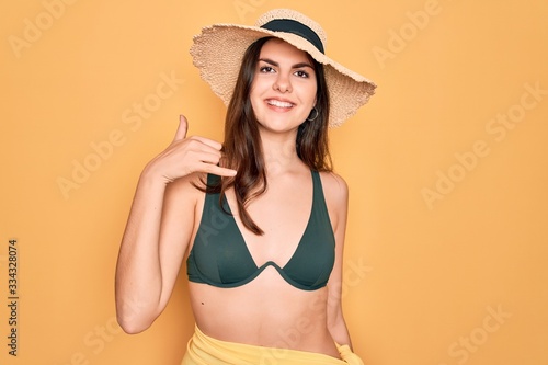 Young beautiful girl wearing swimwear bikini and summer sun hat over yellow background smiling doing phone gesture with hand and fingers like talking on the telephone. Communicating concepts.