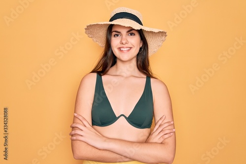 Young beautiful girl wearing swimwear bikini and summer sun hat over yellow background happy face smiling with crossed arms looking at the camera. Positive person.
