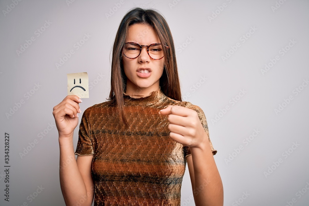 Young beautiful girl holding reminder with sad emotion emoji face over white background annoyed and frustrated shouting with anger, crazy and yelling with raised hand, anger concept Young beautiful girl holding reminder with sad emotion emoji face over white background annoyed and frustrated shouting with anger, crazy and yelling with raised hand, anger concept