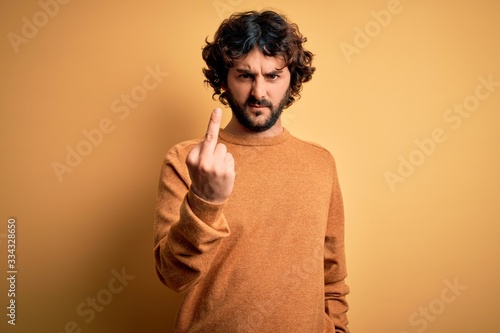 Young handsome man with beard wearing casual sweater standing over yellow background Showing middle finger, impolite and rude fuck off expression
