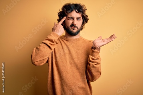 Young handsome man with beard wearing casual sweater standing over yellow background confused and annoyed with open palm showing copy space and pointing finger to forehead. Think about it.