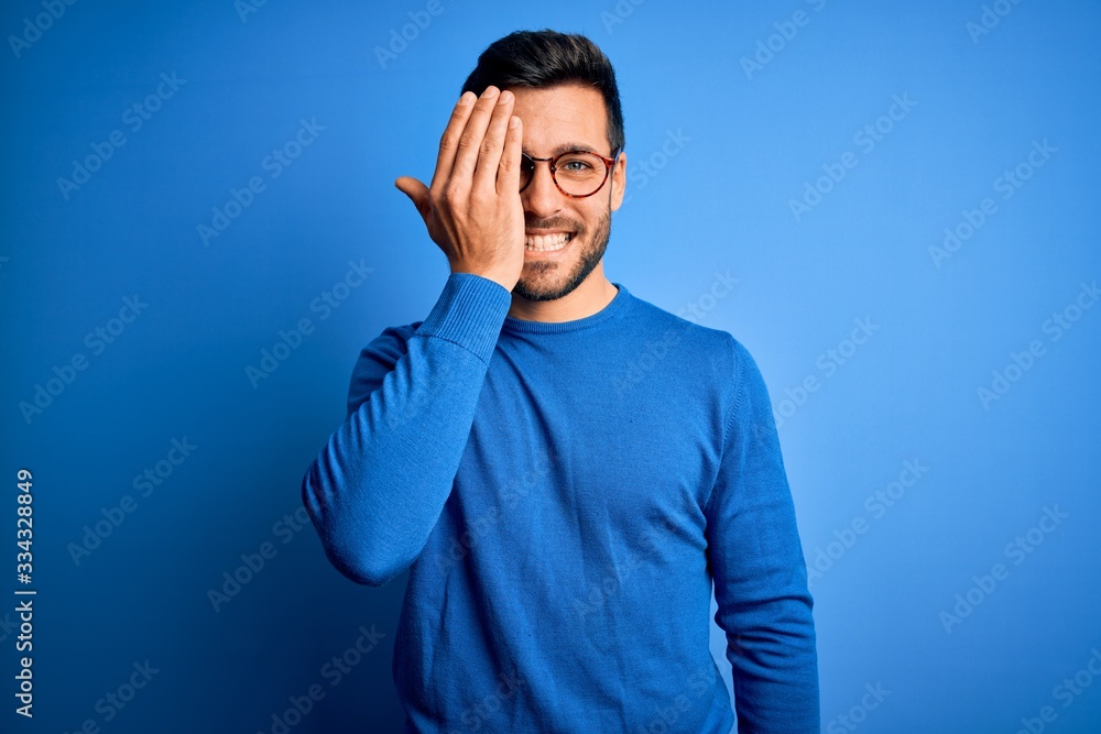Young handsome man with beard wearing casual sweater and glasses over blue background covering one eye with hand, confident smile on face and surprise emotion. Young handsome man with beard wearing casual sweater and glasses over blue background covering one eye with hand, confident smile on face and surprise emotion.