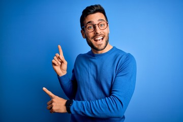 Young handsome man with beard wearing casual sweater and glasses over blue background smiling and looking at the camera pointing with two hands and fingers to the side.