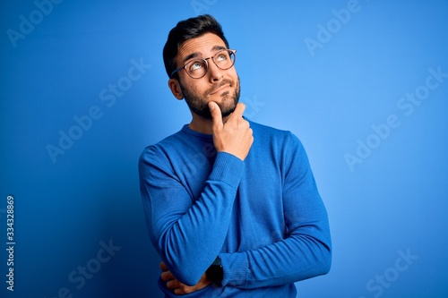 Konstfotografi Young handsome man with beard wearing casual sweater and glasses over blue backg