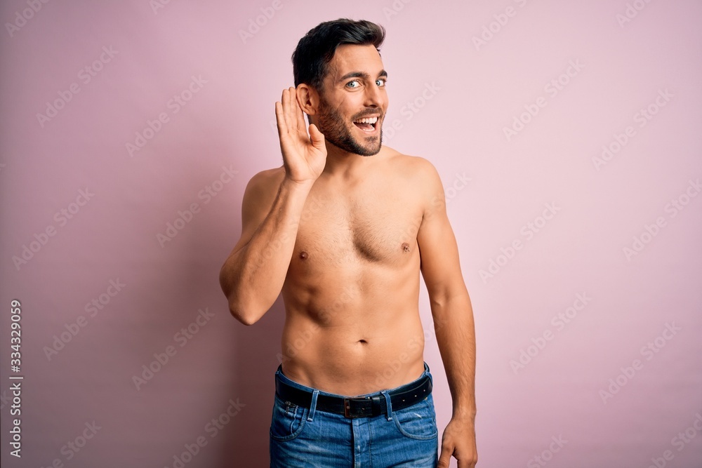 Young handsome strong man with beard shirtless standing over isolated pink background smiling with hand over ear listening an hearing to rumor or gossip. Deafness concept.