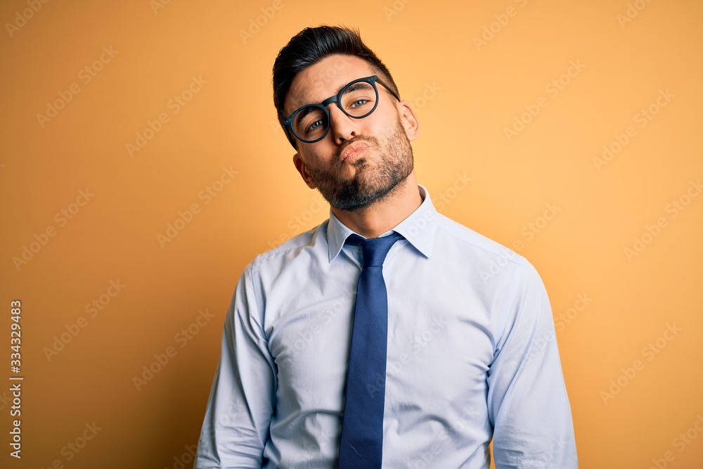 Young handsome businessman wearing tie and glasses standing over yellow background looking at the camera blowing a kiss on air being lovely and sexy. Love expression. Young handsome businessman wearing tie and glasses standing over yellow background looking at the camera blowing a kiss on air being lovely and sexy. Love expression.