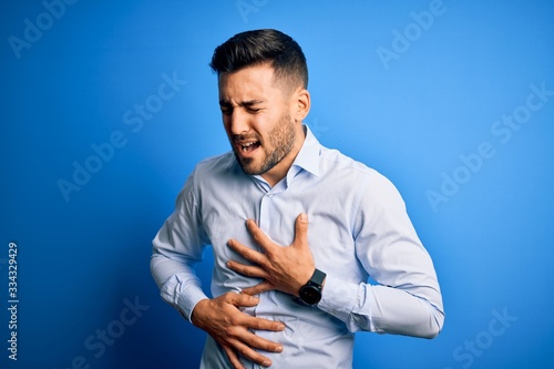 Young handsome man wearing elegant shirt standing over isolated blue background with hand on stomach because nausea, painful disease feeling unwell. Ache concept.
