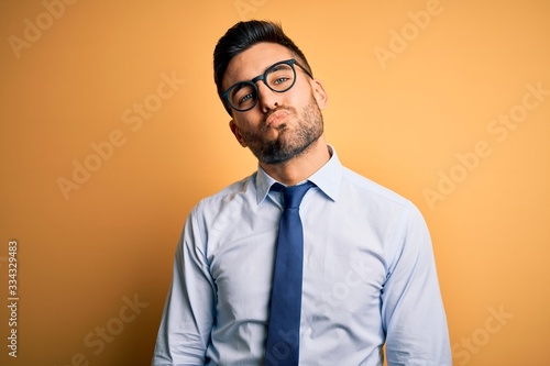 Young handsome businessman wearing tie and glasses standing over yellow background looking at the camera blowing a kiss on air being lovely and sexy. Love expression.