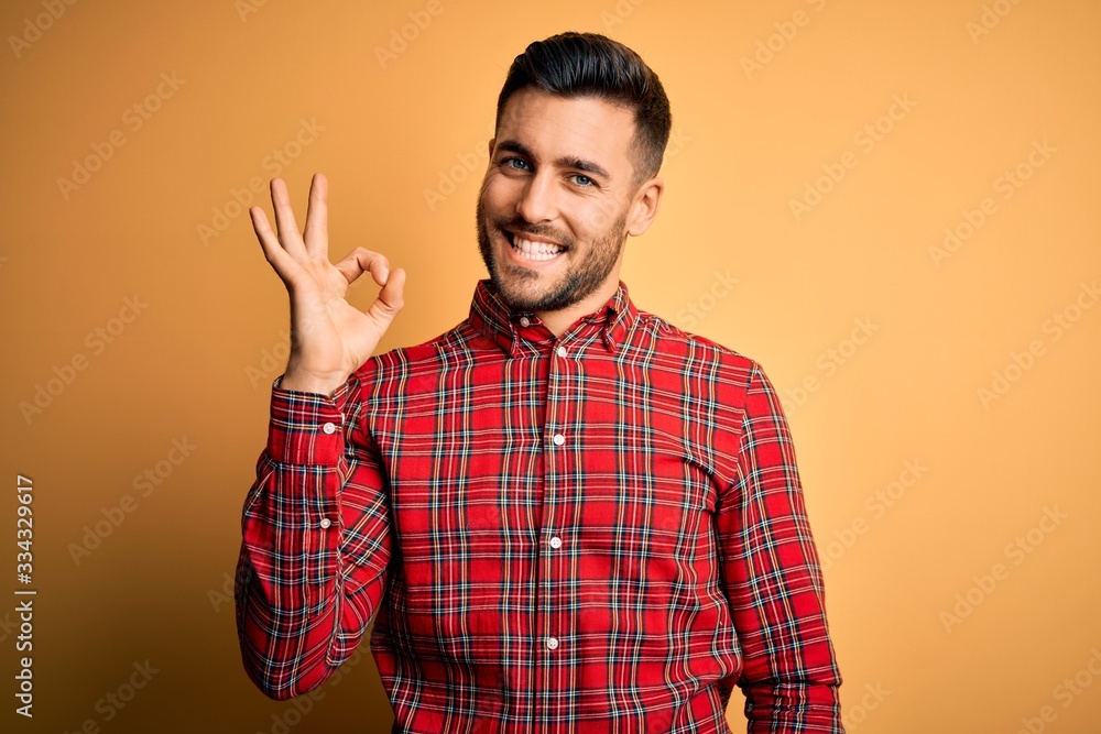 Young handsome man wearing casual shirt standing over isolated yellow background smiling positive doing ok sign with hand and fingers. Successful expression. Young handsome man wearing casual shirt standing over isolated yellow background smiling positive doing ok sign with hand and fingers. Successful expression.