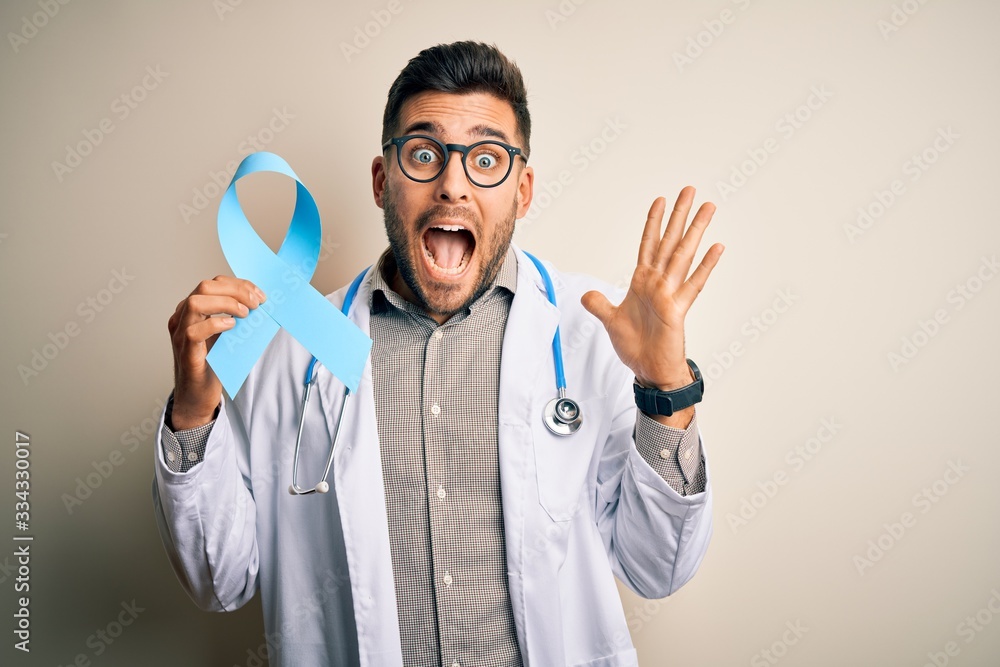 Young professional doctor man holding colon awareness blue ribbon over isolated background very happy and excited, winner expression celebrating victory screaming with big smile and raised hands Young professional doctor man holding colon awareness blue ribbon over isolated background very happy and excited, winner expression celebrating victory screaming with big smile and raised hands