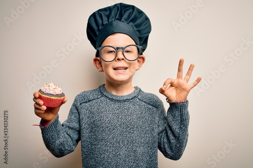 Young little caucasian cook kid wearing chef uniform and hat cooking cupcake doing ok sign with fingers, excellent symbol photo