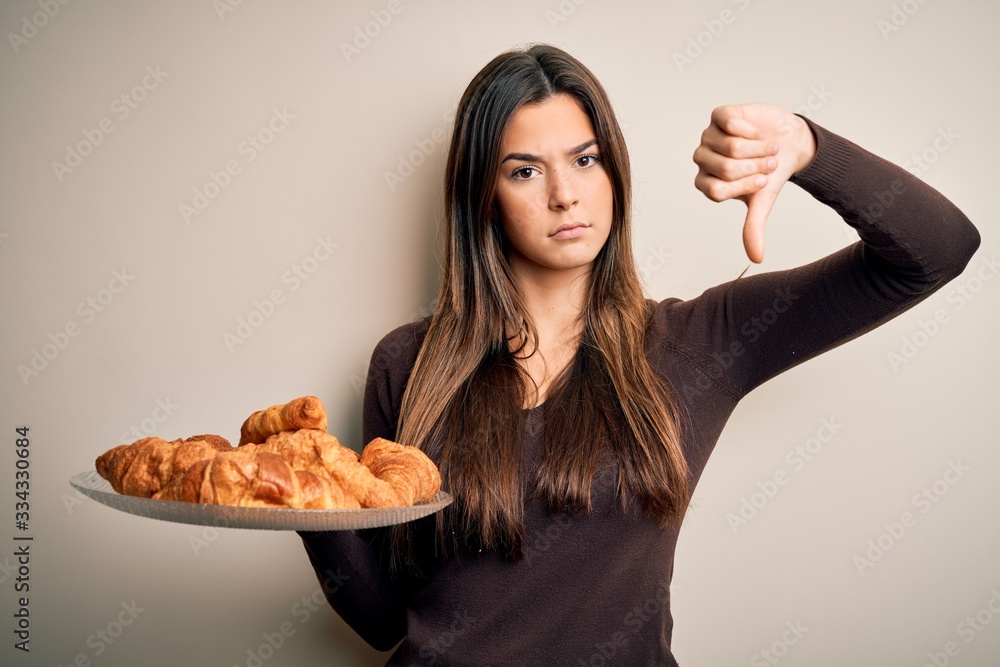 Young beautiful girl holding plate with sweet croissants for breakfast over white background with angry face, negative sign showing dislike with thumbs down, rejection concept
