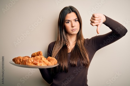 Young beautiful girl holding plate with sweet croissants for breakfast over white background with angry face  negative sign showing dislike with thumbs down  rejection concept