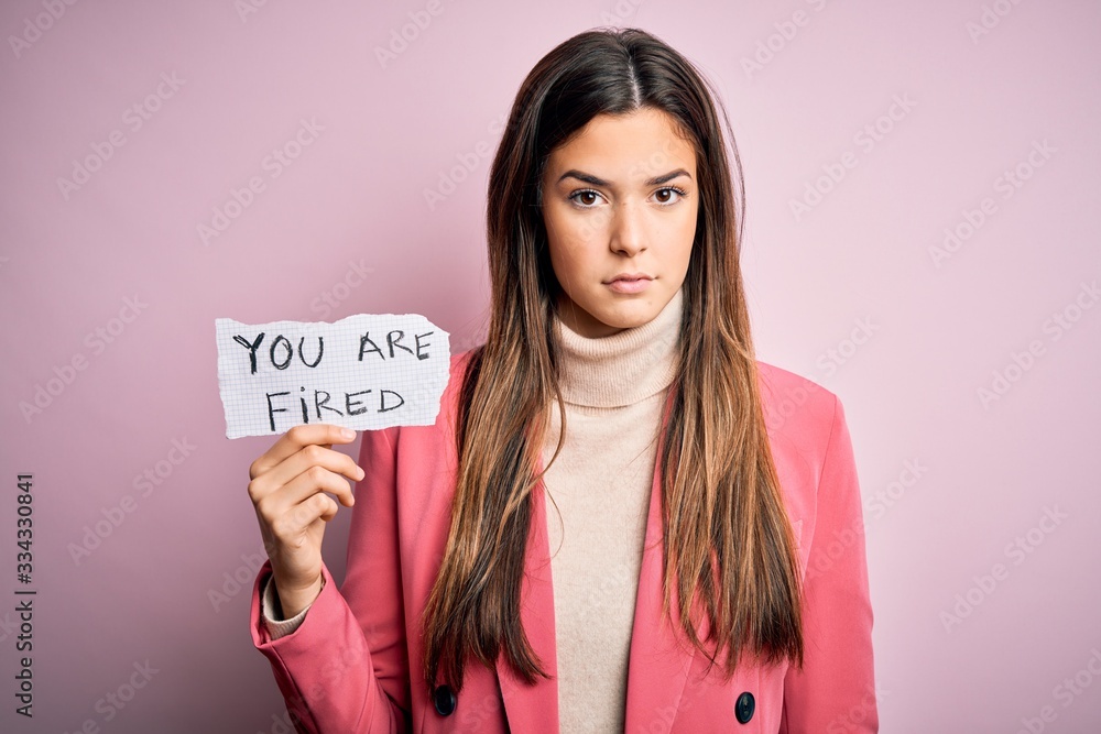 Young beautiful girl holding paper with you are fired message over isolated pink background with a confident expression on smart face thinking serious Young beautiful girl holding paper with you are fired message over isolated pink background with a confident expression on smart face thinking serious