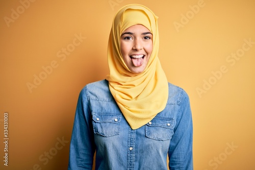 Young beautiful girl wearing muslim hijab standing over isolated yellow background sticking tongue out happy with funny expression. Emotion concept. © Krakenimages.com