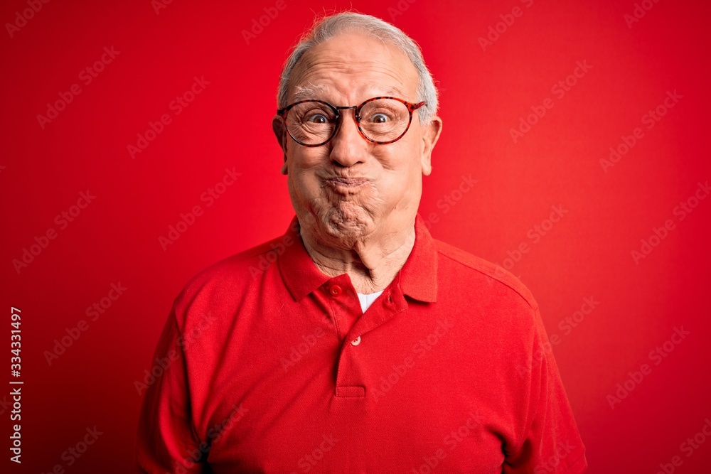 Obraz premium Grey haired senior man wearing glasses and casual t-shirt over red background puffing cheeks with funny face. Mouth inflated with air, crazy expression.