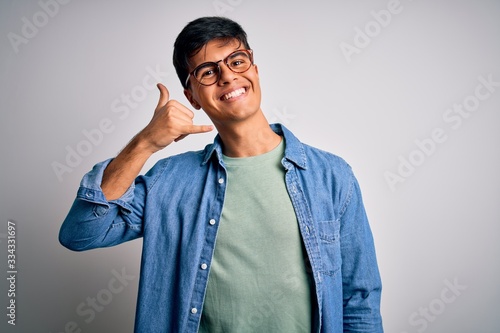 Young handsome man wearing casual shirt and glasses over isolated white background smiling doing phone gesture with hand and fingers like talking on the telephone. Communicating concepts.